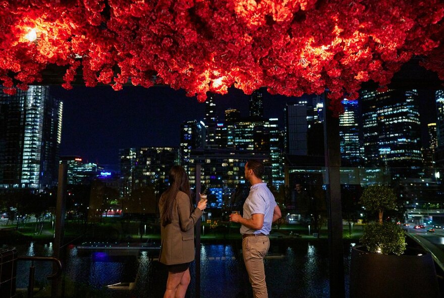 A couple sharing a drink under red flowers looking at the city skyline at night.