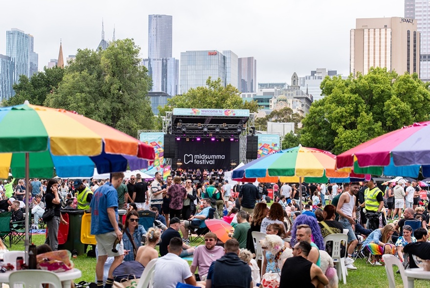 A large crowd at an outdoor stage with rainbow picnic umbrellas in the foreground and the Melbourne city skyline in the background.