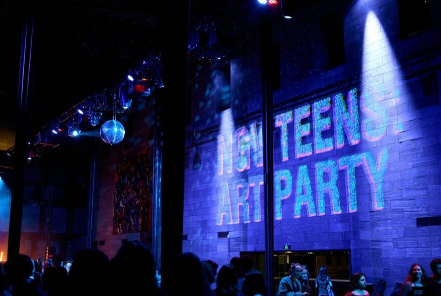 A dark venue room with a projection on the wall saying NGV TEENS ART PARTY, a disco ball hanging from the roof. 