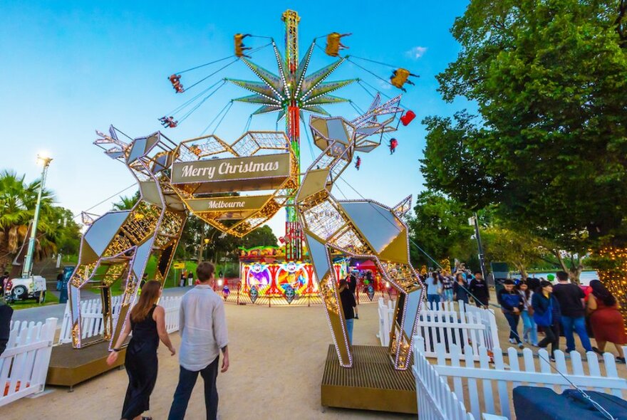 A couple walking past a deer decoration towards carnival rides, trees and crowds just before dusk.