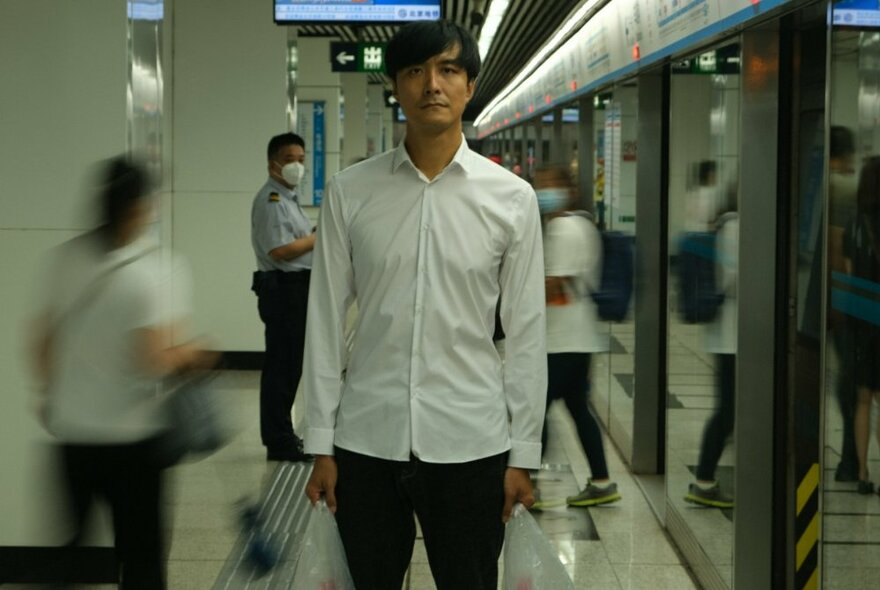 Man wearing a white shirt and dark trousers, standing upright facing the viewer in a white tiled arcade, with figures in motion behind and around him.