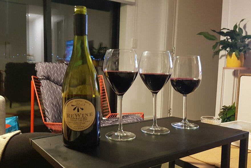 A ReWine's refillable glass bottle and three glasses half-filled with red wine on a coffee table in a domestic lounge setting.