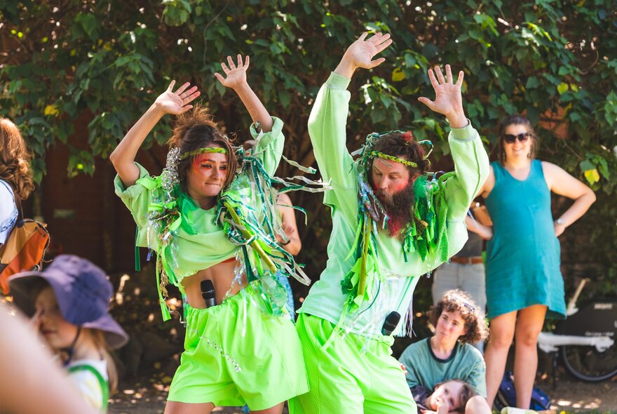 Two people dressed in lime green shorts and t-shirts dancing outdoors in a public space with people smiling and watching.