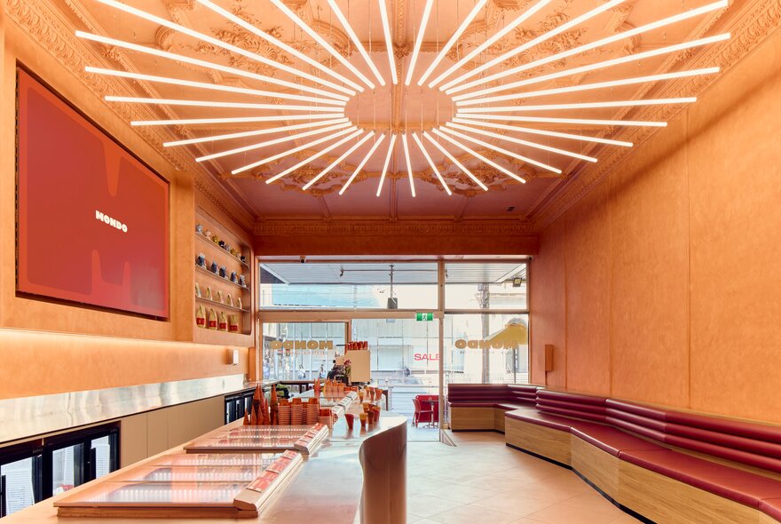 A long interior of a retail shop with warm orange walls, a white counter, red bench seating, and a striking ceiling light fixture made of many white neon tubes radiating from a central point.