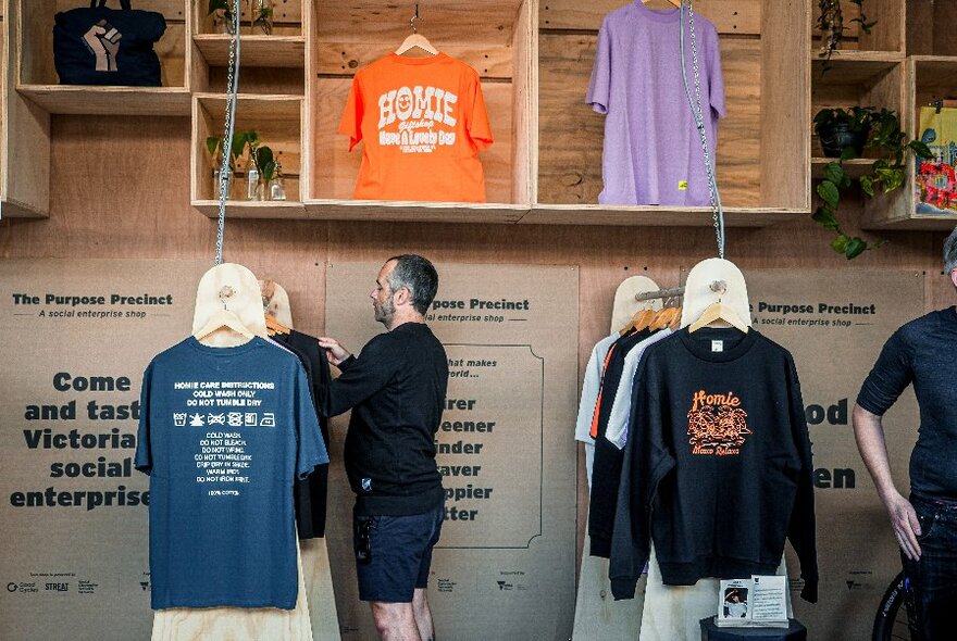 Man seen from side, arranging t-shirts on a rack, with another closing rack to his right, and wooden display cases above.