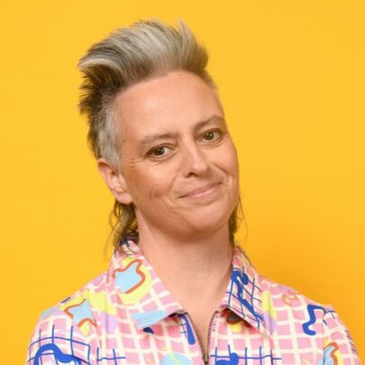 Portrait of comedian Geraldine Hickey, wearing a colourful shirt and standing in front of a yellow background.