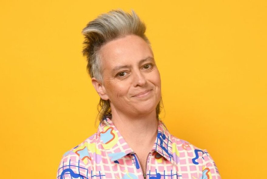Portrait of comedian Geraldine Hickey, wearing a colourful shirt and standing in front of a yellow background.