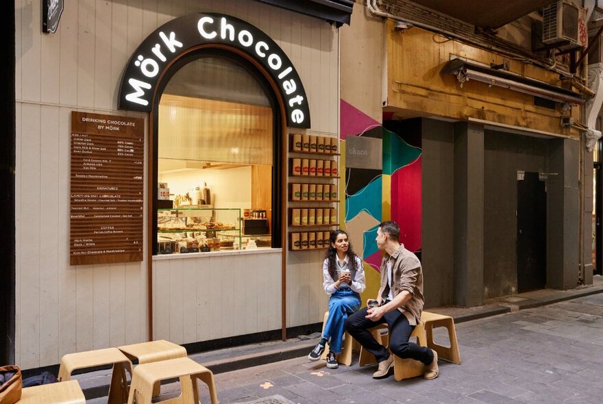 Small chocolate store with serving window in laneway, two people sitting on stools out the front.