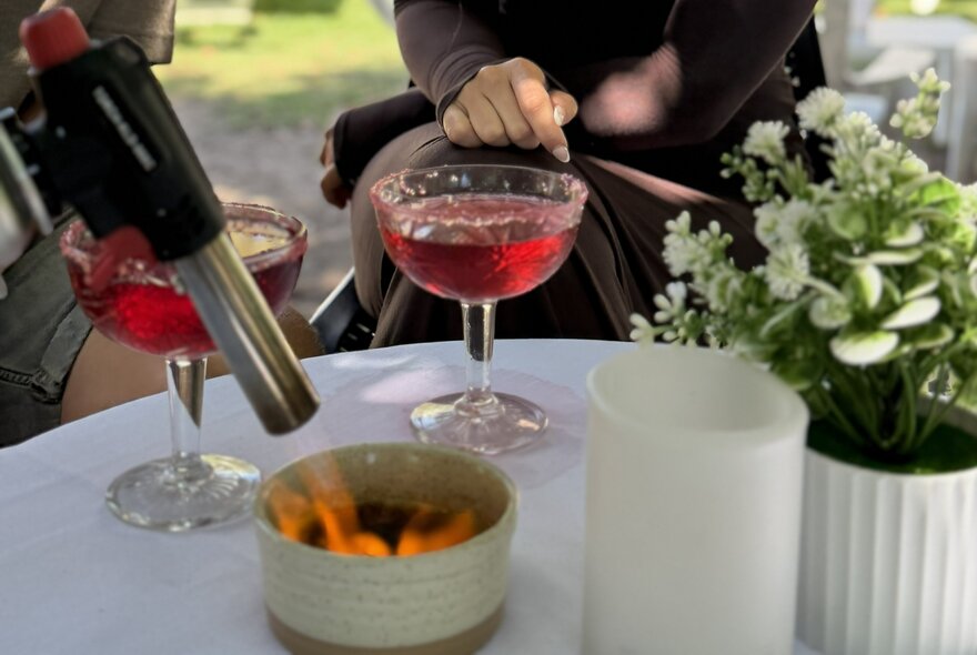 A person holds a kitchen torch over a creme brulee, with two red cocktails, and white potted flowers on a white table.