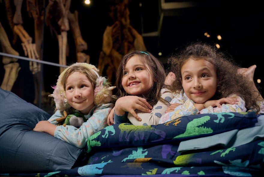Three little girls smiling under a doona, dinosaur skeleton legs visible in the background. 