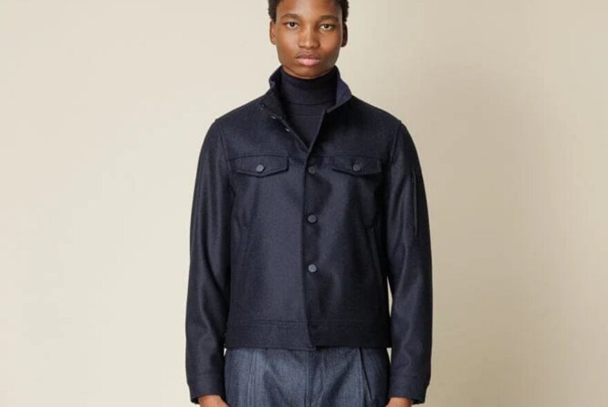 A male model wearing a tailored dark navy blue bomber style aviator jacket over a dark turtle neck jumper.