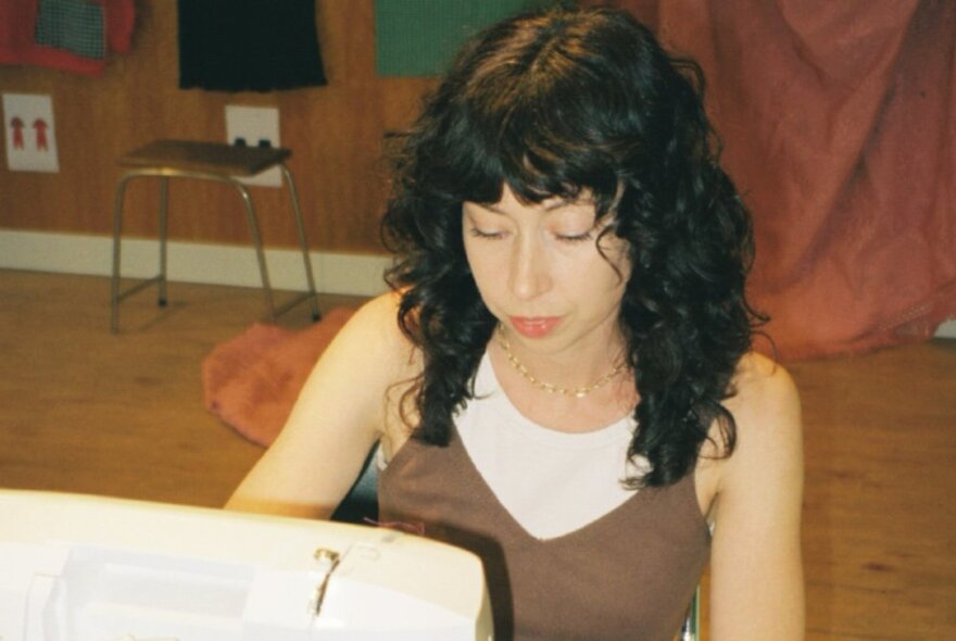 A woman with her eyes looking downwards, with curled dark hair, seated and working at a sewing machine.