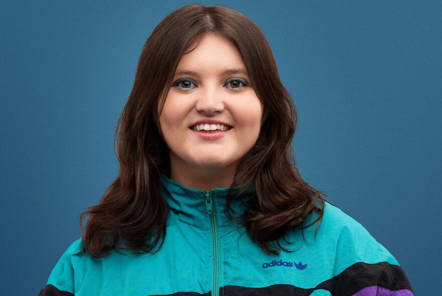 A smiling young woman wearing an aqua parachute jacket, with her long brown hair over her shoulders.