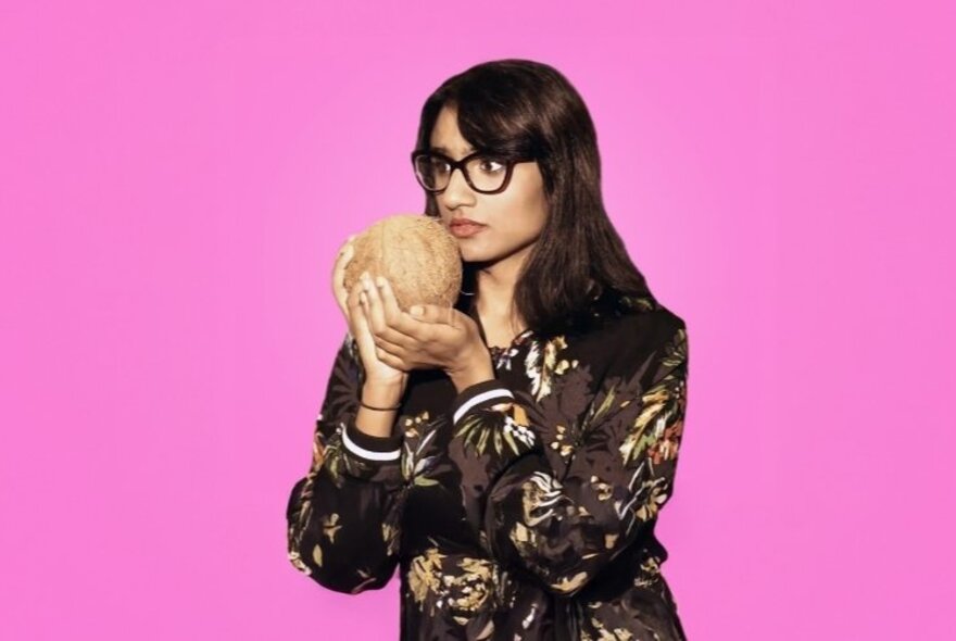 Vidya Rajan, a woman shown in half-bust format, wearing a patterned brown long-sleeve top and holding a whole coconut close to her face, posed against a pink background. 