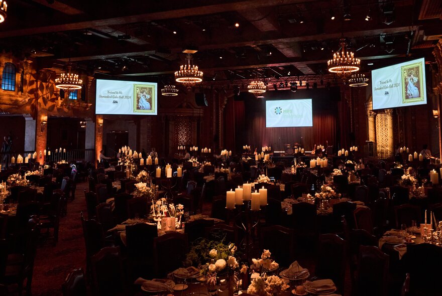 Dimly lit dining venue with candle-lit tables, chandeliers and overhead screens.