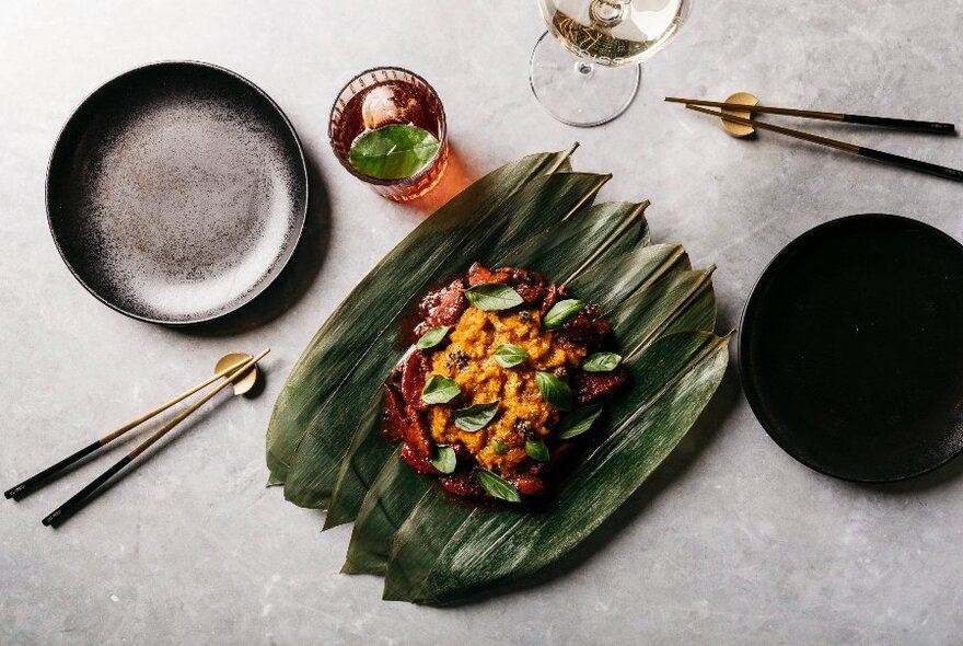 Looking down at a colourful meal presented on green pandan leaves, chopsticks resting on holders.