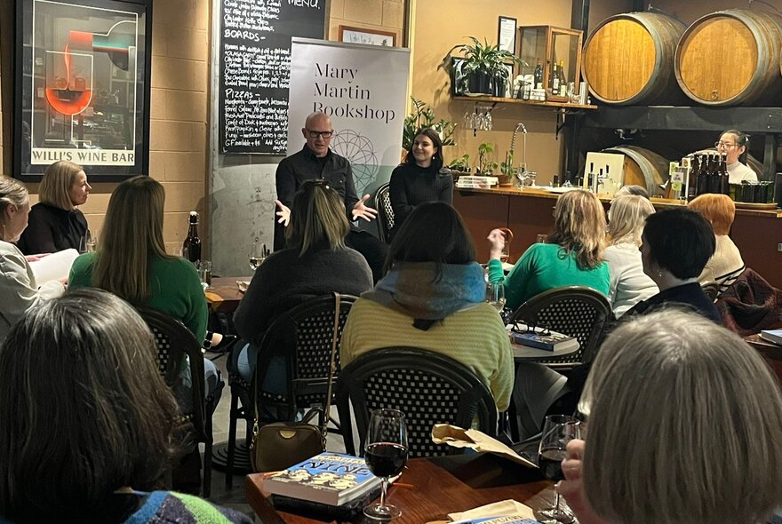 People seated in a wine bar with barrels, listening to two speakers in front of signage.
