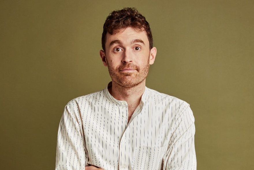 Portrait of comedian Tom Cashman, wearing a slightly questioning look and standing in front of a khaki background.