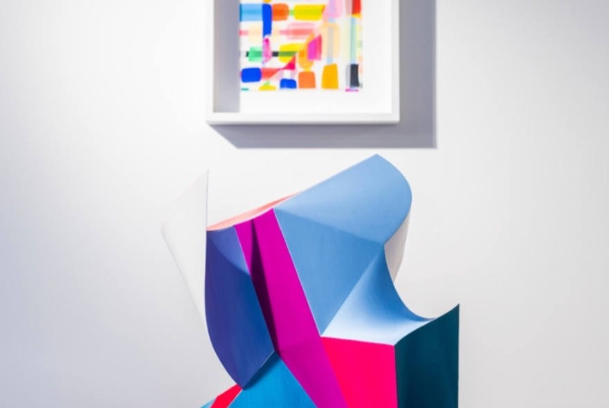 Pink and blue abstract sculpture in front of a white wall and smaller abstract painting.