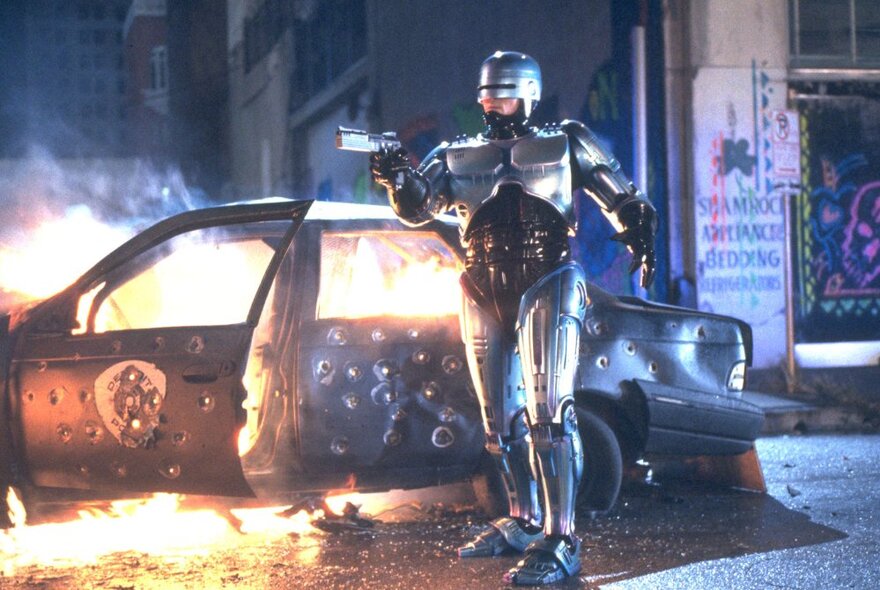 A silver robot with a gun outside a car on fire.