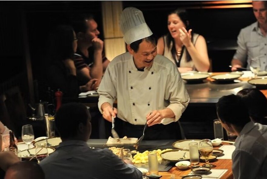 A Japanese Teppanyaki chef cooking at a traditional Teppan grill, with patrons seated at the table which surrounds the grill.