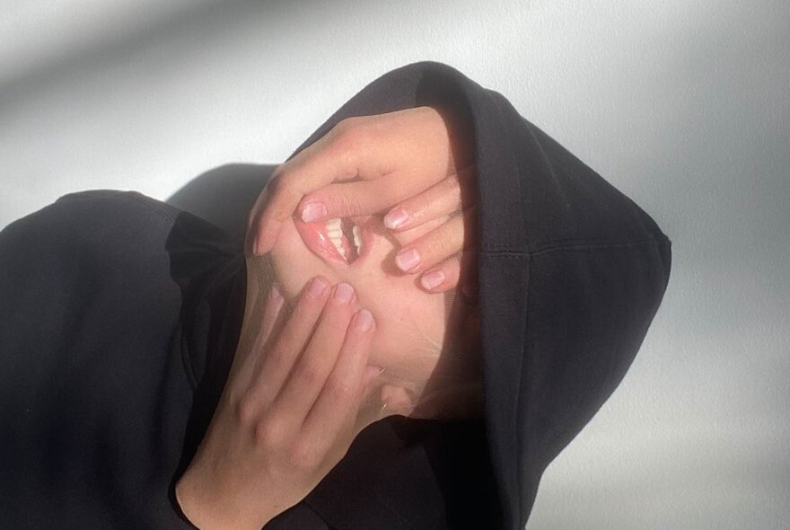 Person's face with three hands half covering it, encased in a sheer stocking, wearing a black hoodie top. 