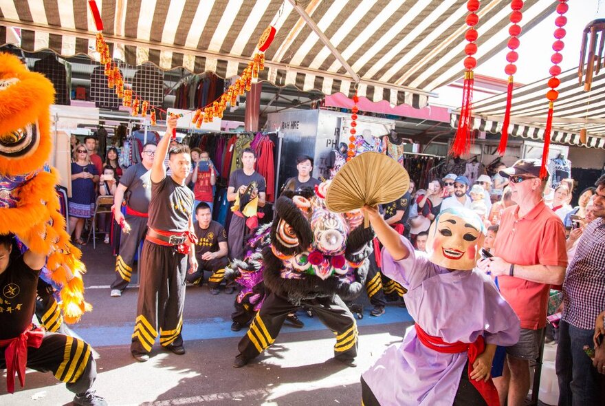 Lion dance performers dancing with traditional outfits under a canopy at Queen Vic Market.