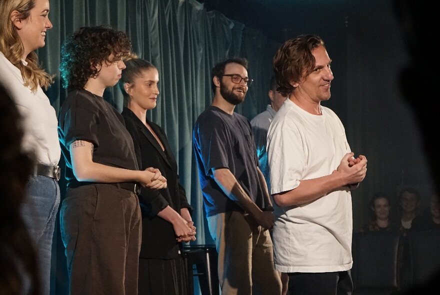 Performers standing in a row on a stage in front of a curtain, with one performer standing slightly in front of the others.
