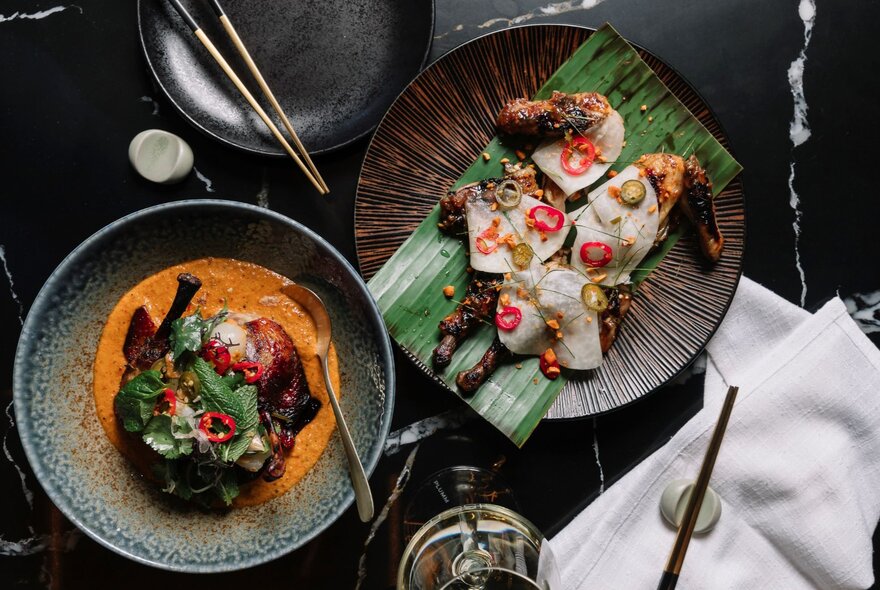 Looking down on two Asian style dishes, one on pandan leaves and another in a red curry sauce. 