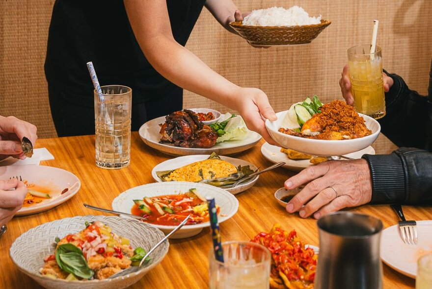 A person in black handing out plates of Indonesian food in a restaurant setting.