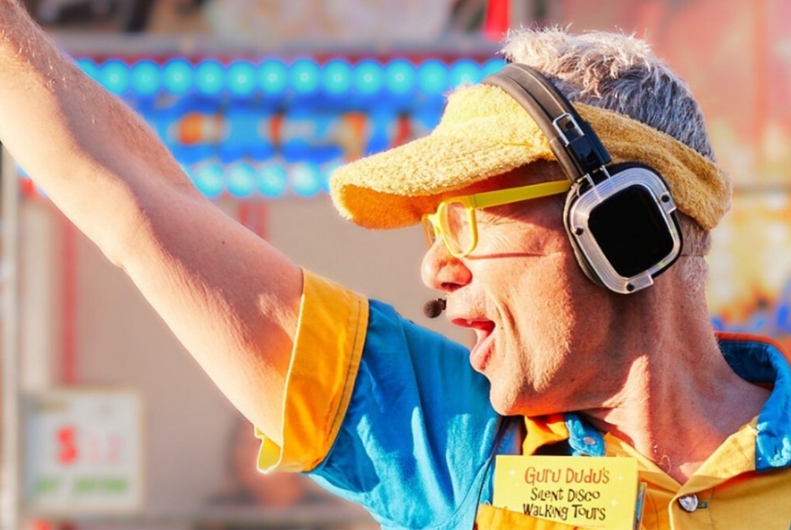 A man in profile wearing an orange sun visor and headphones over his ears, his arm pointing up.