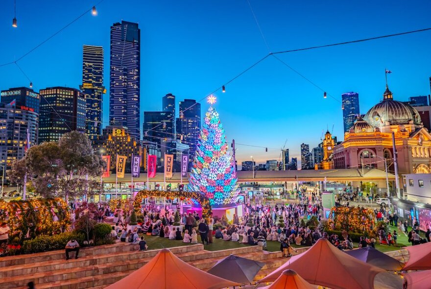 Crowds gathered around Federation Square's Christmas tree and decorations at dusk, with cafe umbrellas, Flinders Street Station and city buildings. 