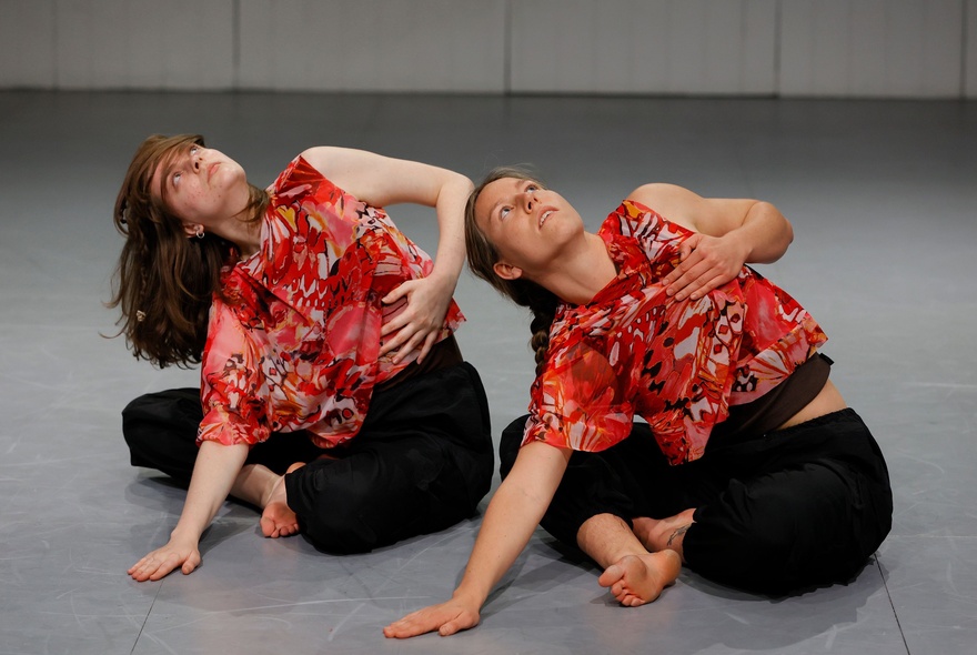 Two dancers dressed in red patterned top and black pants performing on a floor, seated, with their upper bodies twisting towards the ceiling.