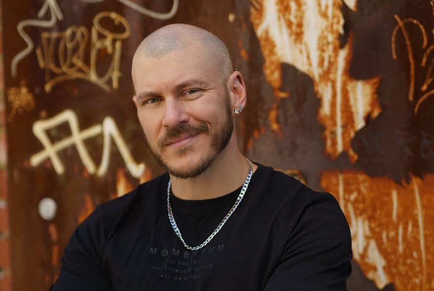A man with a shaved head wearing a black t-shirt and silver chain, smiling, posing in front of a graffitied wall.