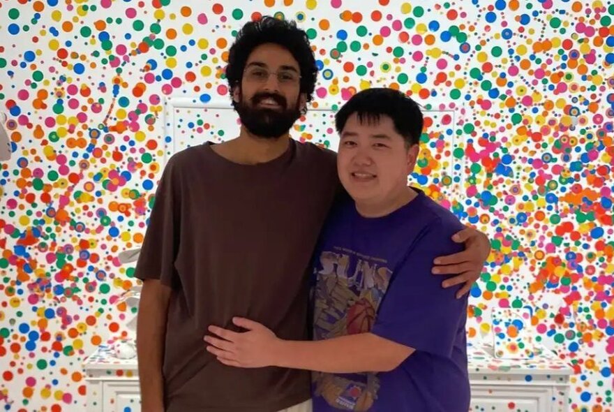 Two friends hugging in front of a colourful paint-speckled wall.