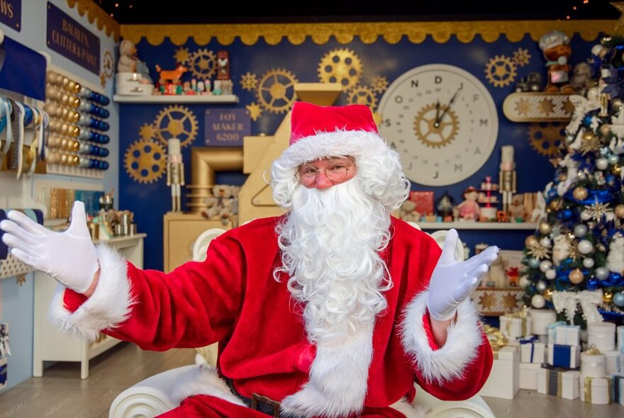 Santa with his gloved white hands outstretched, seated in front of a an room decked out with Christmas decorations, and a small Christmas tree.