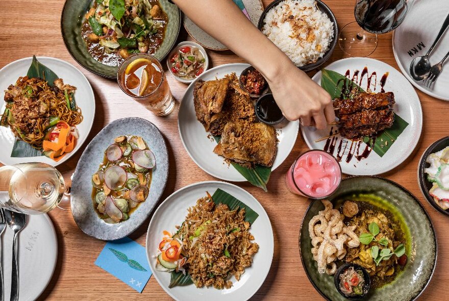 Overhead view of several patters of food, and drinks on a table, with a hand reaching across to pick up a satay stick.