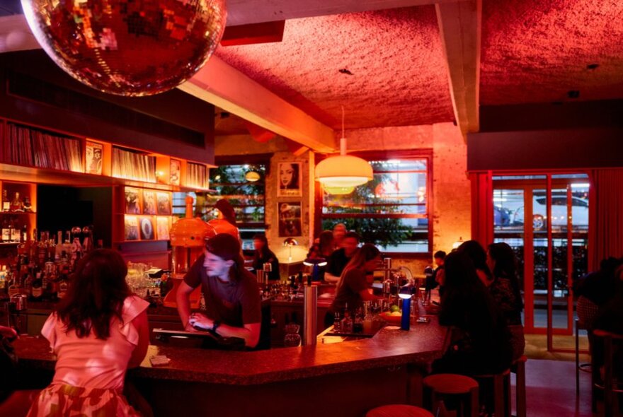 Cocktail bar interior with mirror ball, red walls and bar staff speaking to seated customers at the bar.