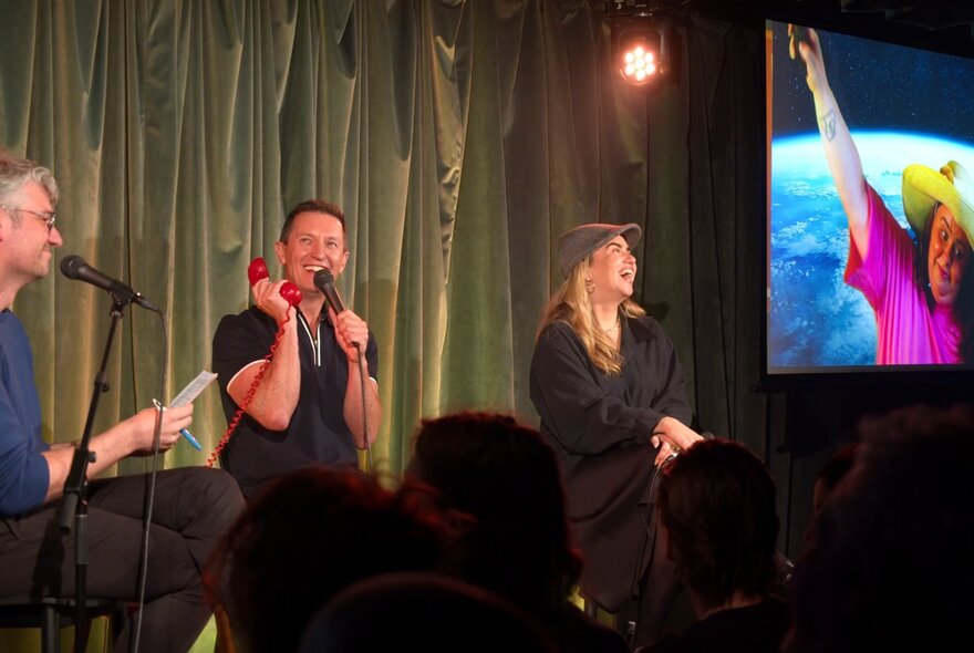A group of improv comedians seated on stools  on a stage in front of an audience, with a large monitor to their left.