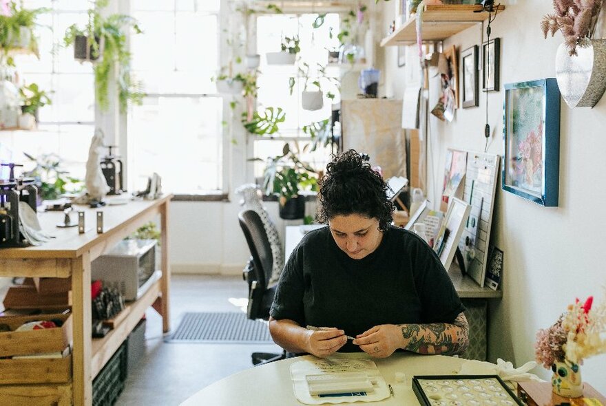 Women with dark hair working on jewellery at a table, light and plant-filled studio at rear.