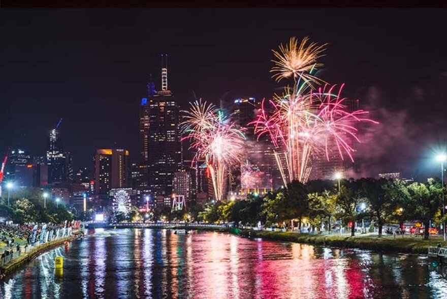 Nighttime fireworks display on the banks of Melbourne's Yarra River, with city skyscrapers in the background.