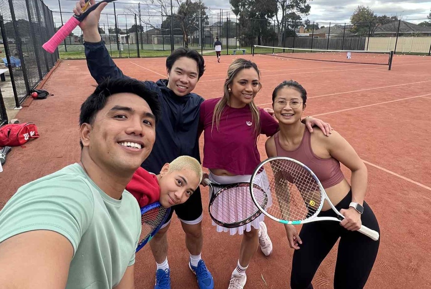 A man taking a group selfie of his four friends on a tennis court. 