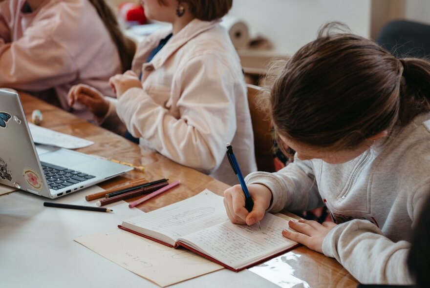 Young teenagers writing in notebooks or on laptops in a creative writing workshop in a studio setting.