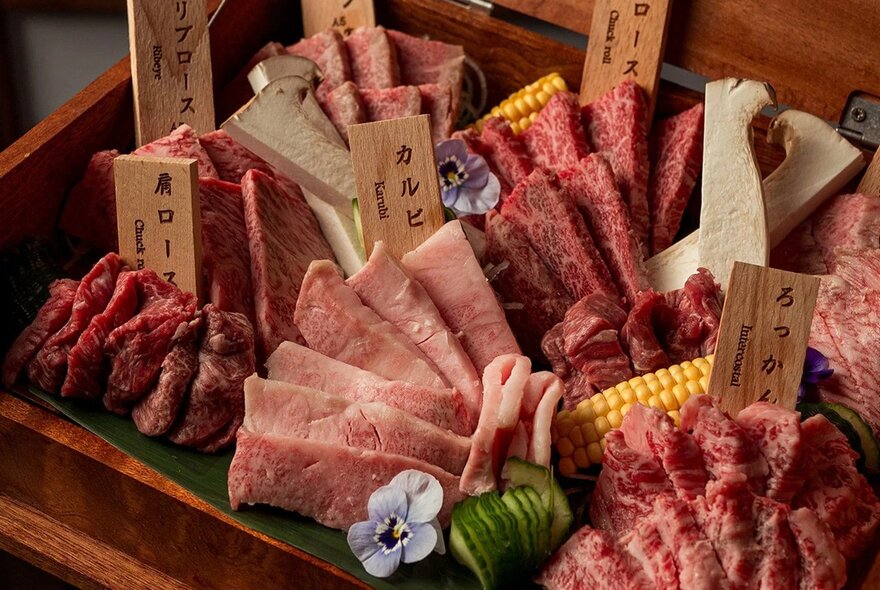 Wooden box of raw Japanese ingredients including meat and corn.