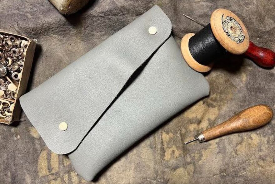 A grey leather purse made from repurposed leather, on a concrete table with leather-working tools.