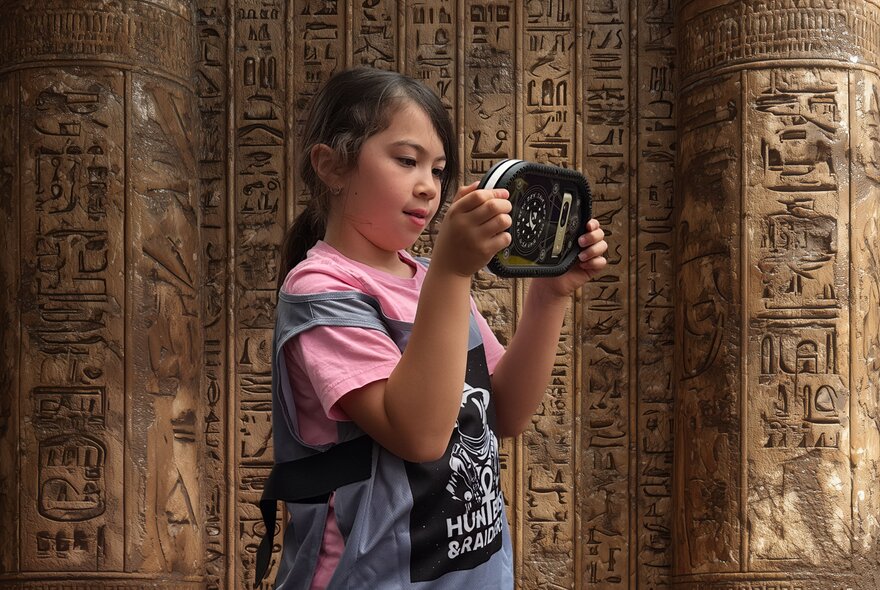 A child looking at a digital device in front of a wall decorated with Ancient Egyptian Hieroglyphics.