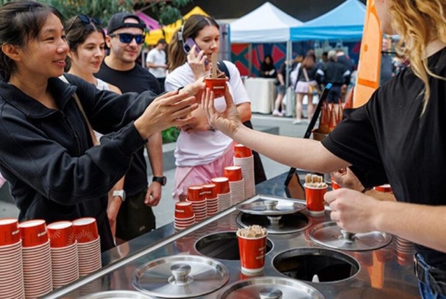 A person handing a small drink or dessert cup from an outdoor market stall or cart to a customer.