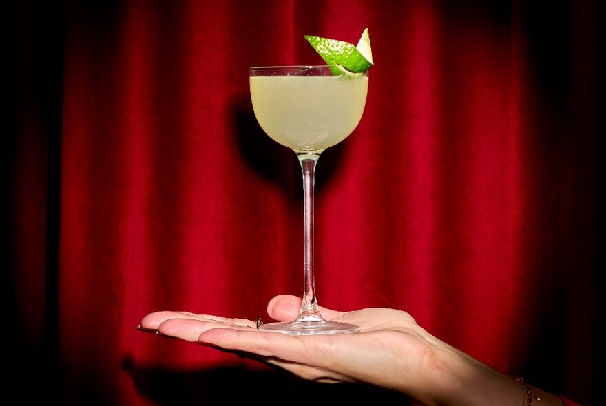 An open-palm of a hand balancing a cocktail against a red curtain backdrop.