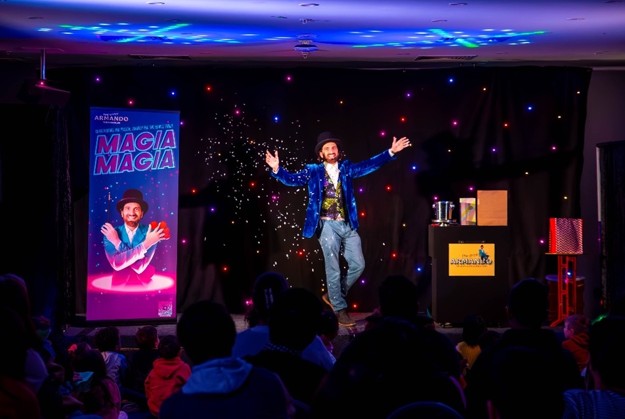 Magician performing on stage, wearing a blue jacket with his arms outstretched.