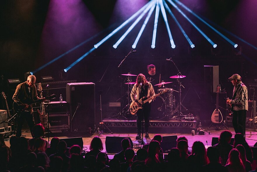 A band performs live on a stage with purple lighting and bright blue light beams shining outwards from the back.